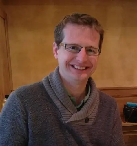 Jordan Raddick looking forward and smiling, wearing glasses and a brown sweater.