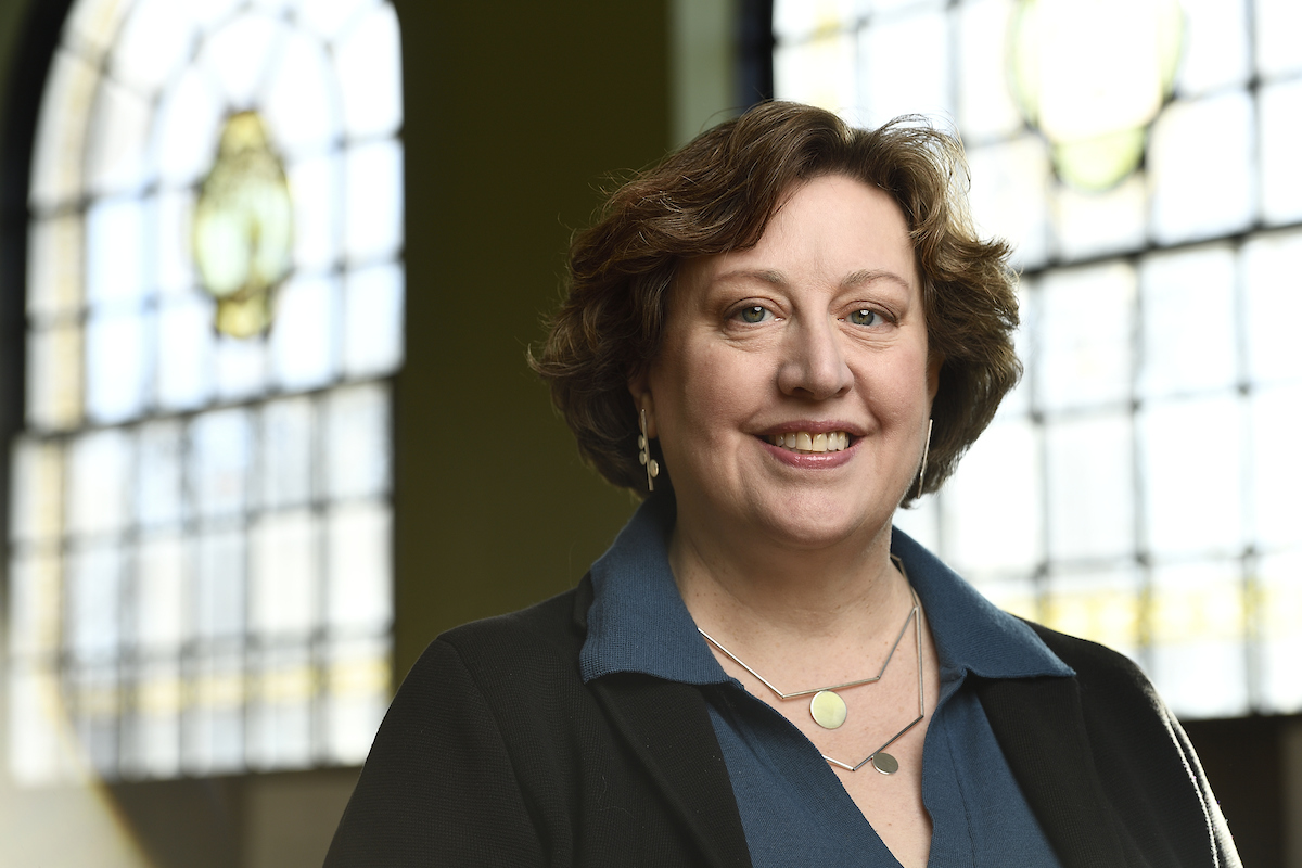 Dean Elisabeth Long in front of stained glass windows, looking forward and smiling, wearing blue collared shirt with black jacket.